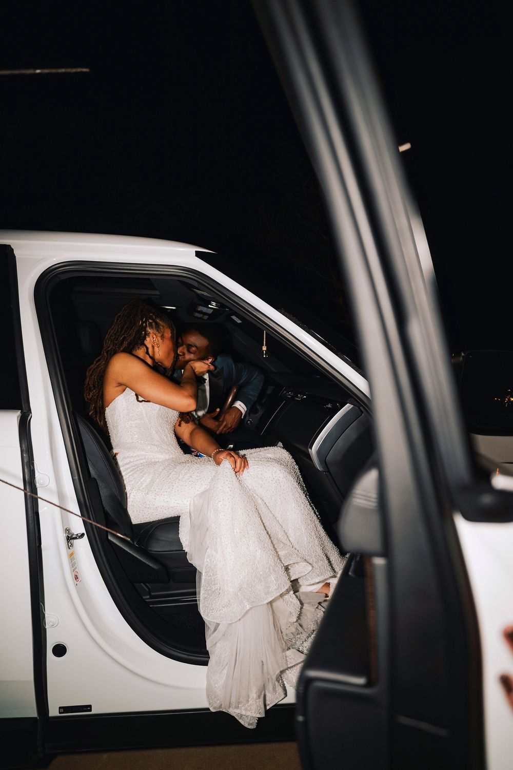 Bride in white gown sits on groom's lap sharing a kiss inside a white truck at night.
