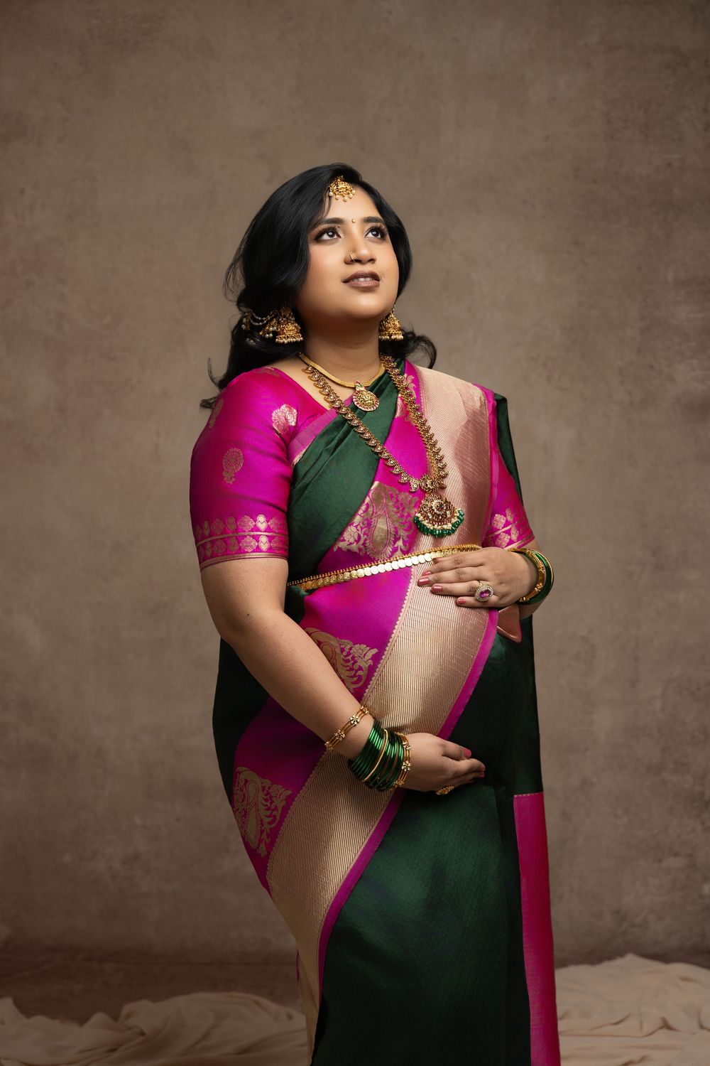 Woman in green and pink silk saree with gold jewelry poses gracefully in studio portrait.
