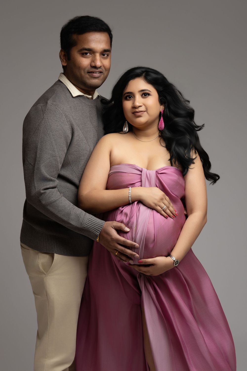 A pregnant woman in a pink gown poses with her partner in a studio maternity photoshoot.