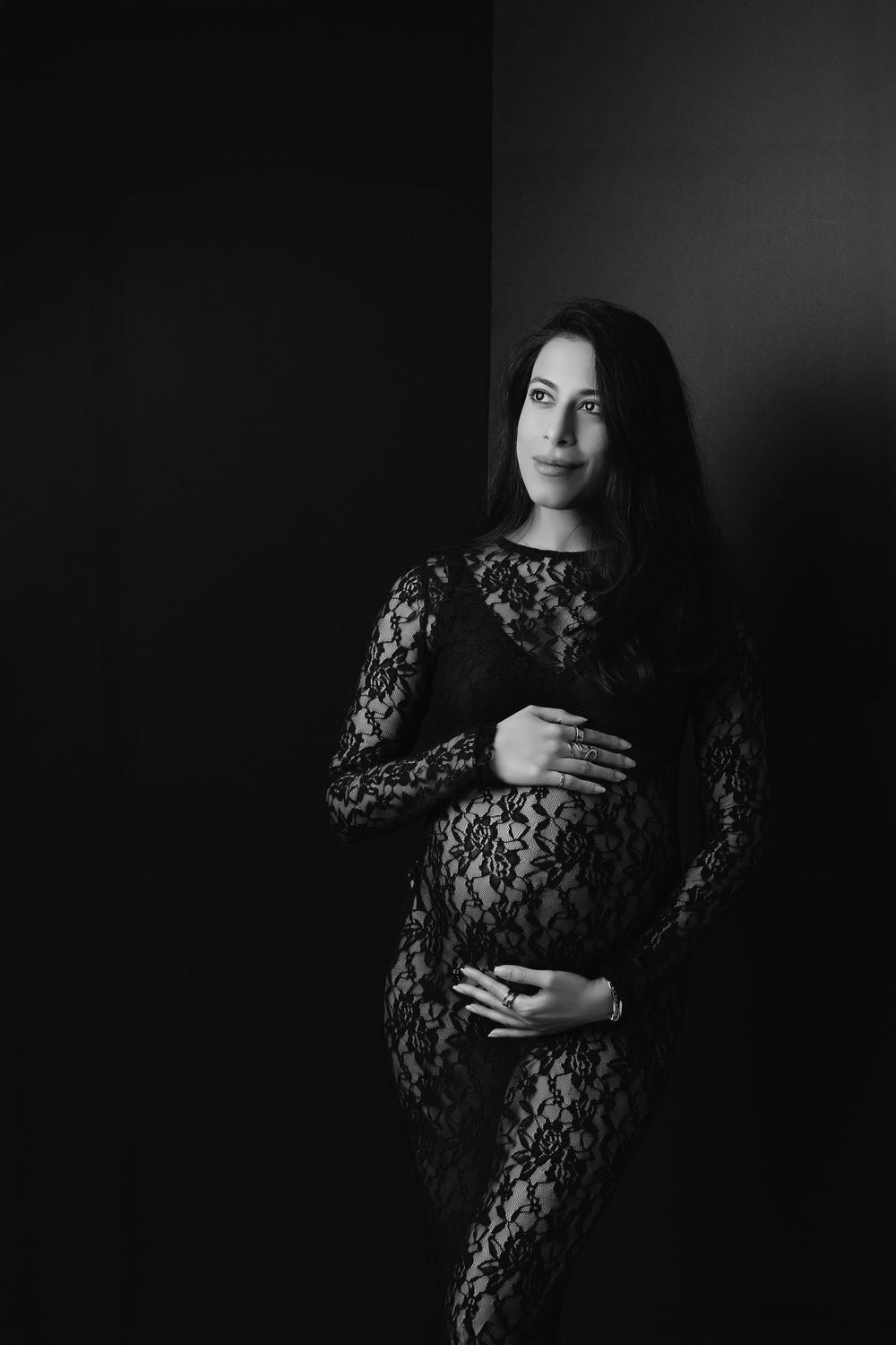 Black and white portrait of pregnant woman in lace dress cradling her baby bump against dark background.