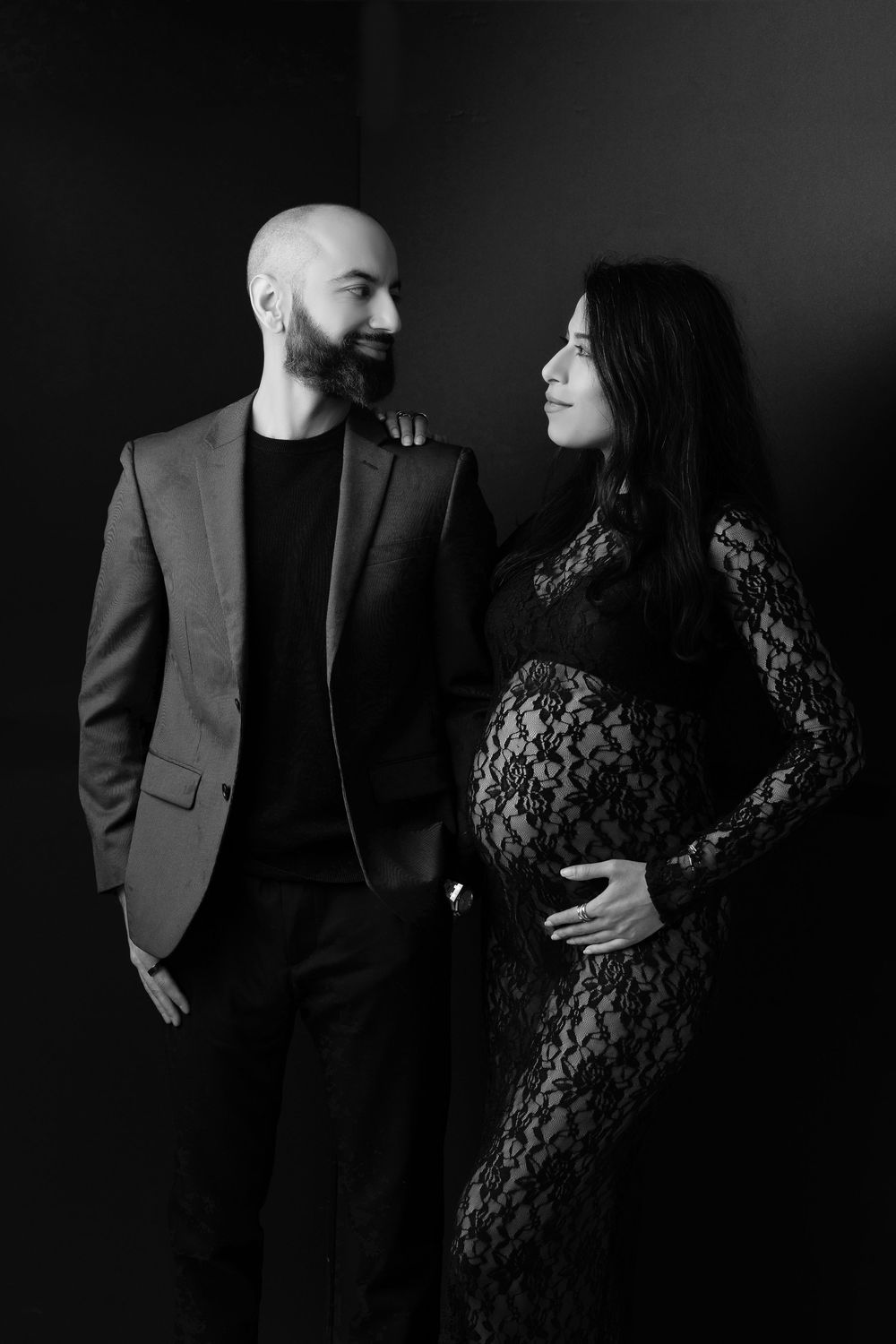Black and white maternity photo of a bald bearded man in a suit smiling at his pregnant partner in a lace dress.