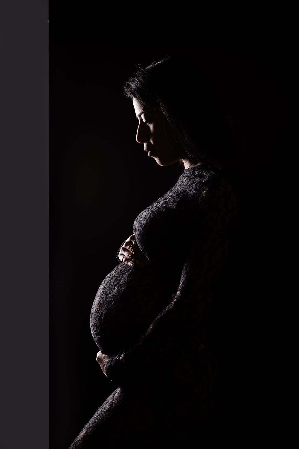 Pregnant woman in dark studio, cradling baby bump in lace dress, dramatic silhouette profile portrait.