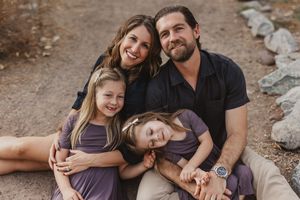 Happy family of four posing outdoors on a dirt path, parents with two young daughters in purple dresses.