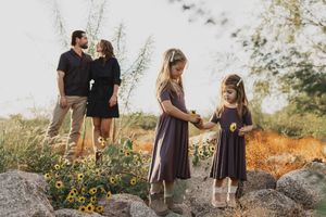 Family photo with two girls holding sunflowers in a field, parents standing together in the background.