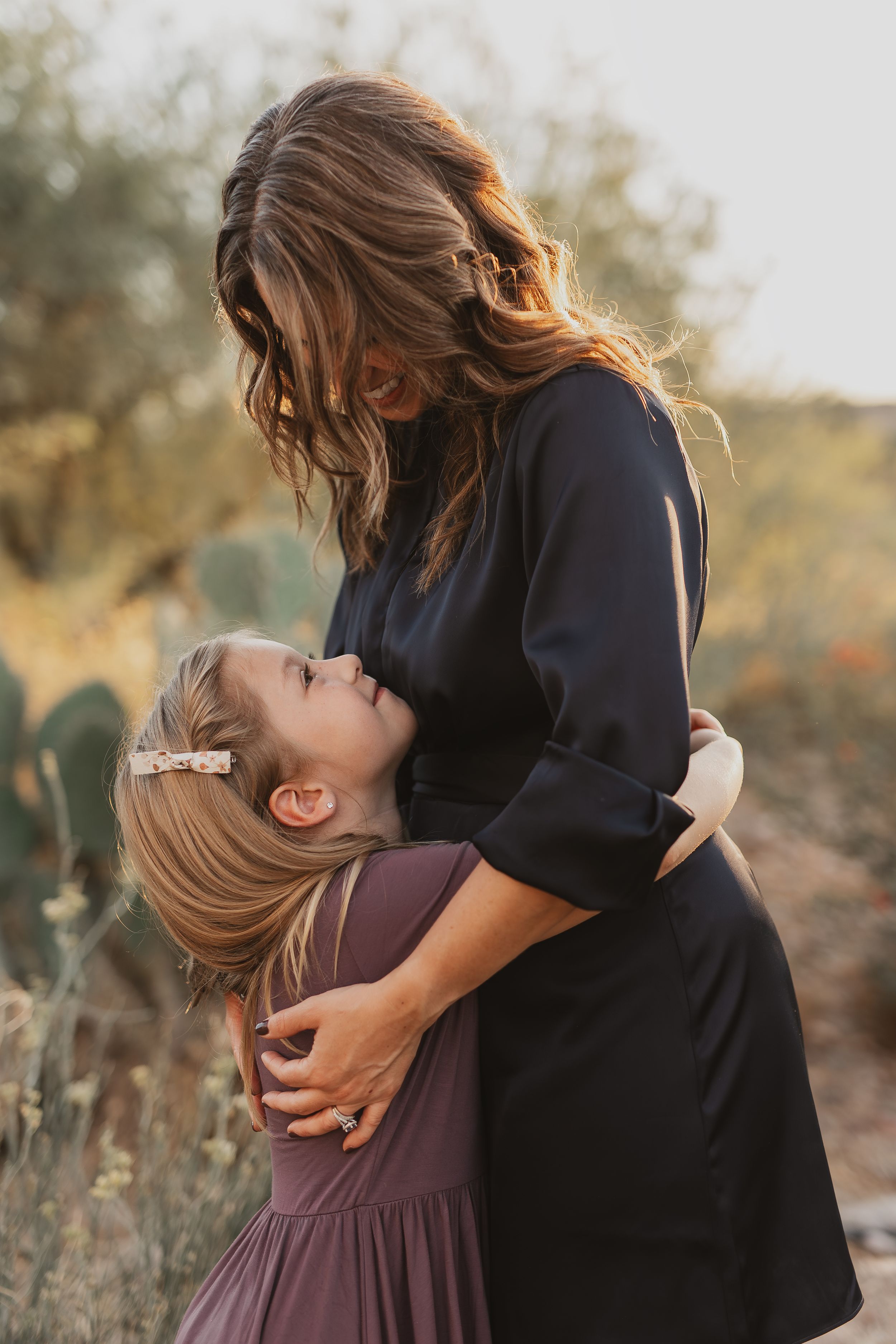A mother in a navy dress tenderly embraces her young daughter in a floral hair clip outdoors at golden hour.
