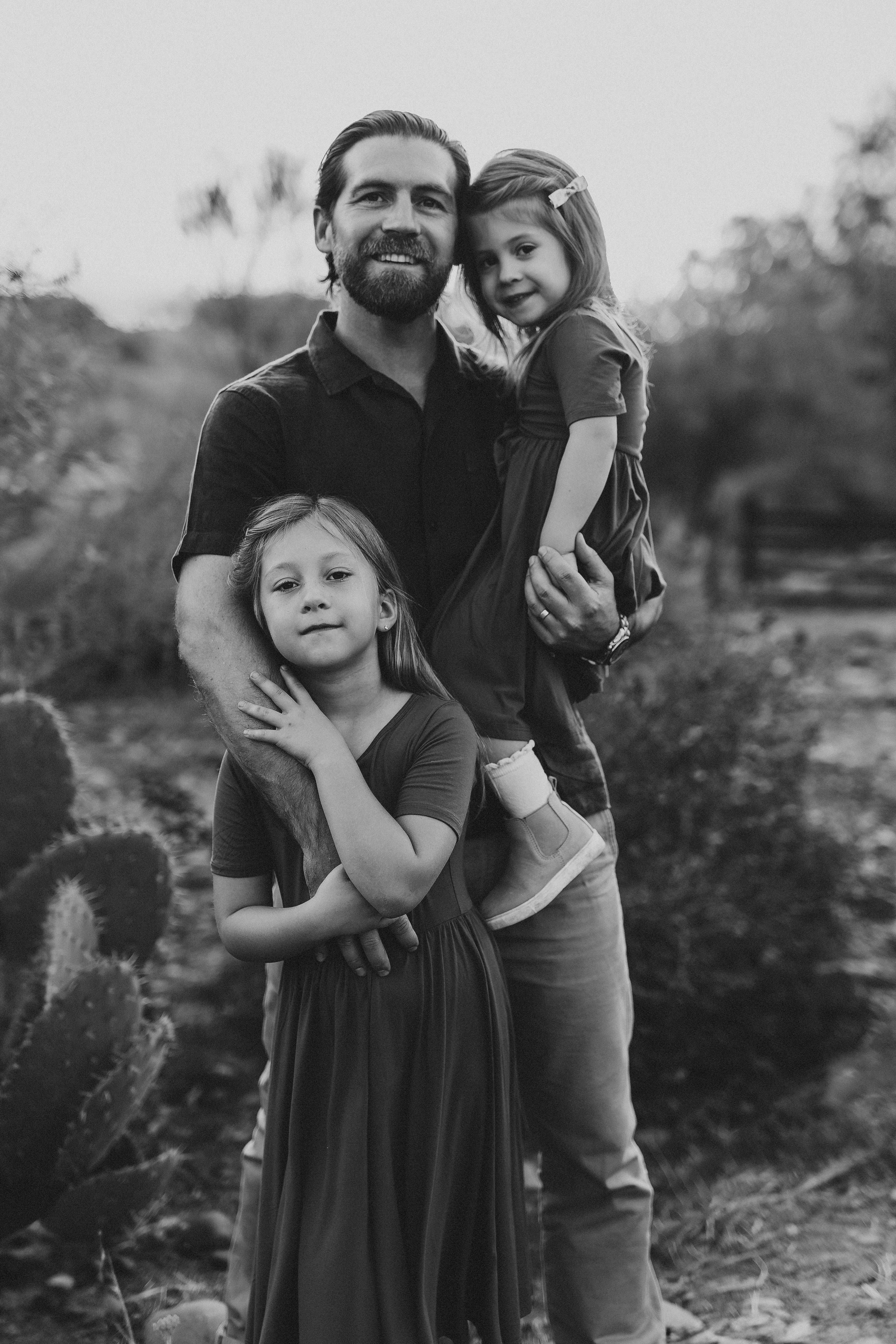 Black and white photo of a smiling father holding two young daughters outdoors among desert cacti.