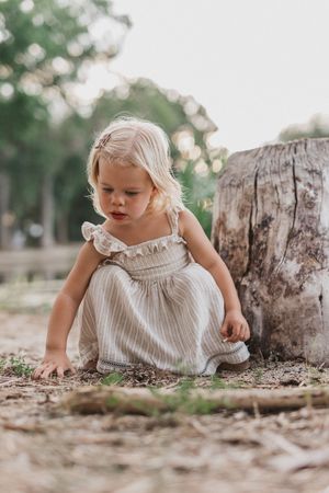 A blonde toddler girl in a striped sundress explores the ground near a tree stump outdoors.