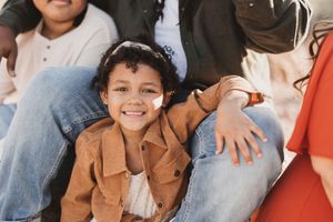 A smiling young child in a brown jacket sits with family members wearing jeans outdoors in a warm, candid portrait.