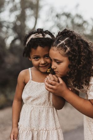 Two curly-haired girls in floral dresses share a sweet moment smelling a small yellow flower outdoors.