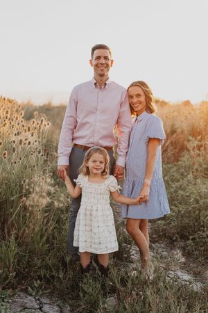 A smiling family of three poses in a golden field at sunset, with parents holding hands with their young daughter in a white dress.