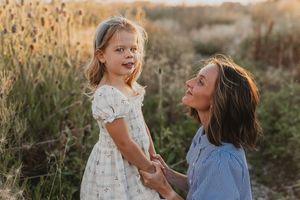 A mother kneels in a golden field holding hands with her young blonde daughter in a white floral dress at sunset.