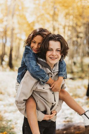 Two smiling teens enjoy a piggyback ride outdoors in an autumn forest with golden yellow leaves.