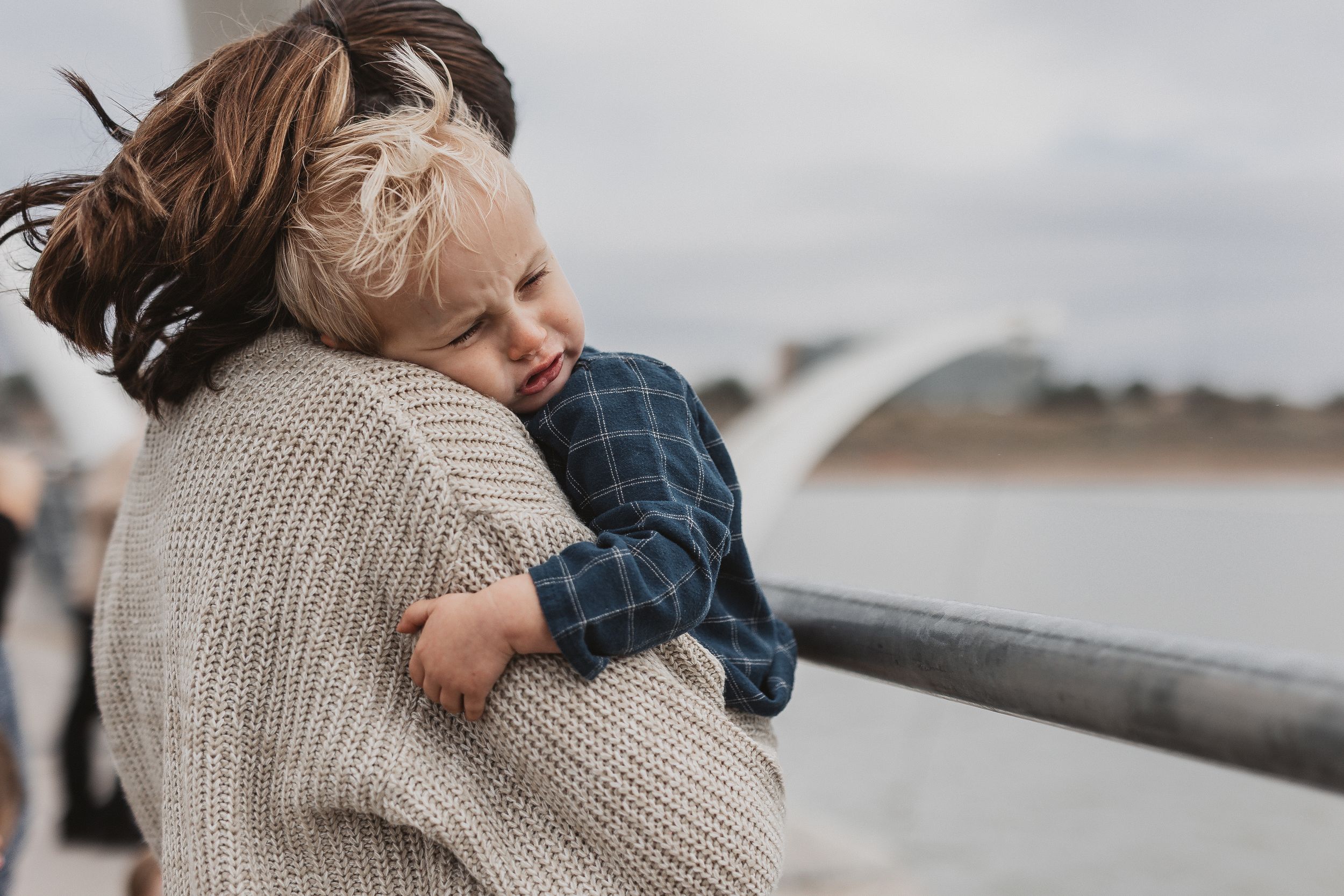 A mother tenderly holds her young blonde toddler on a bridge, both dressed in casual knitwear on a cloudy day.