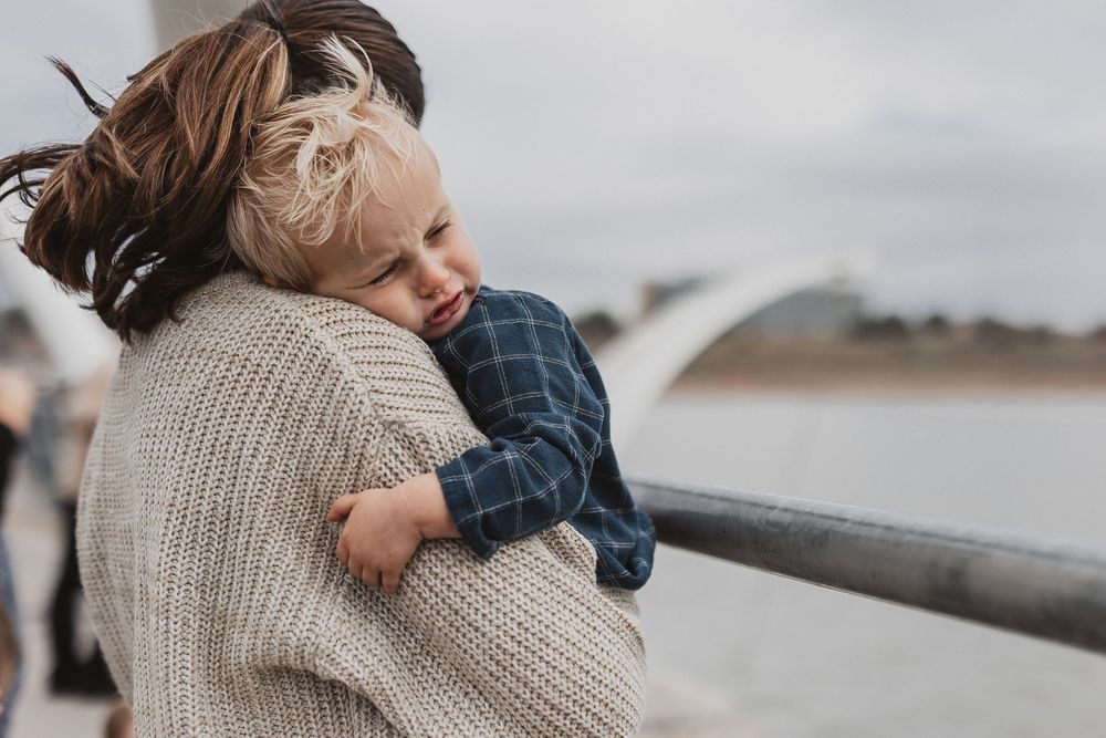 A mother tenderly holds her young blonde toddler on a bridge, both dressed in casual knitwear on a cloudy day.