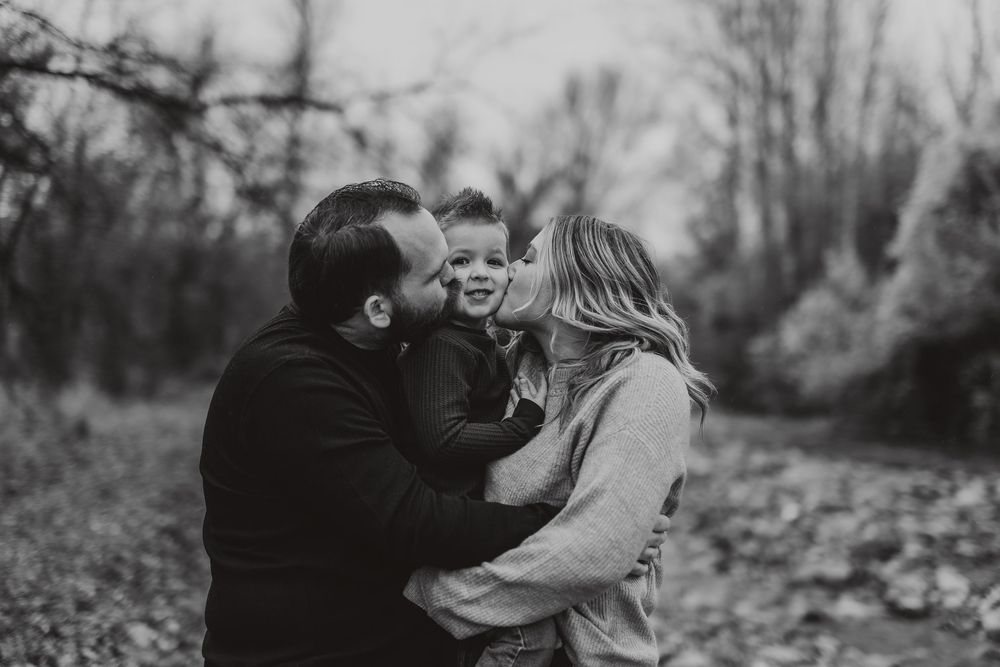 Black and white photo of parents kissing their young child on the cheeks outdoors in a wooded setting.