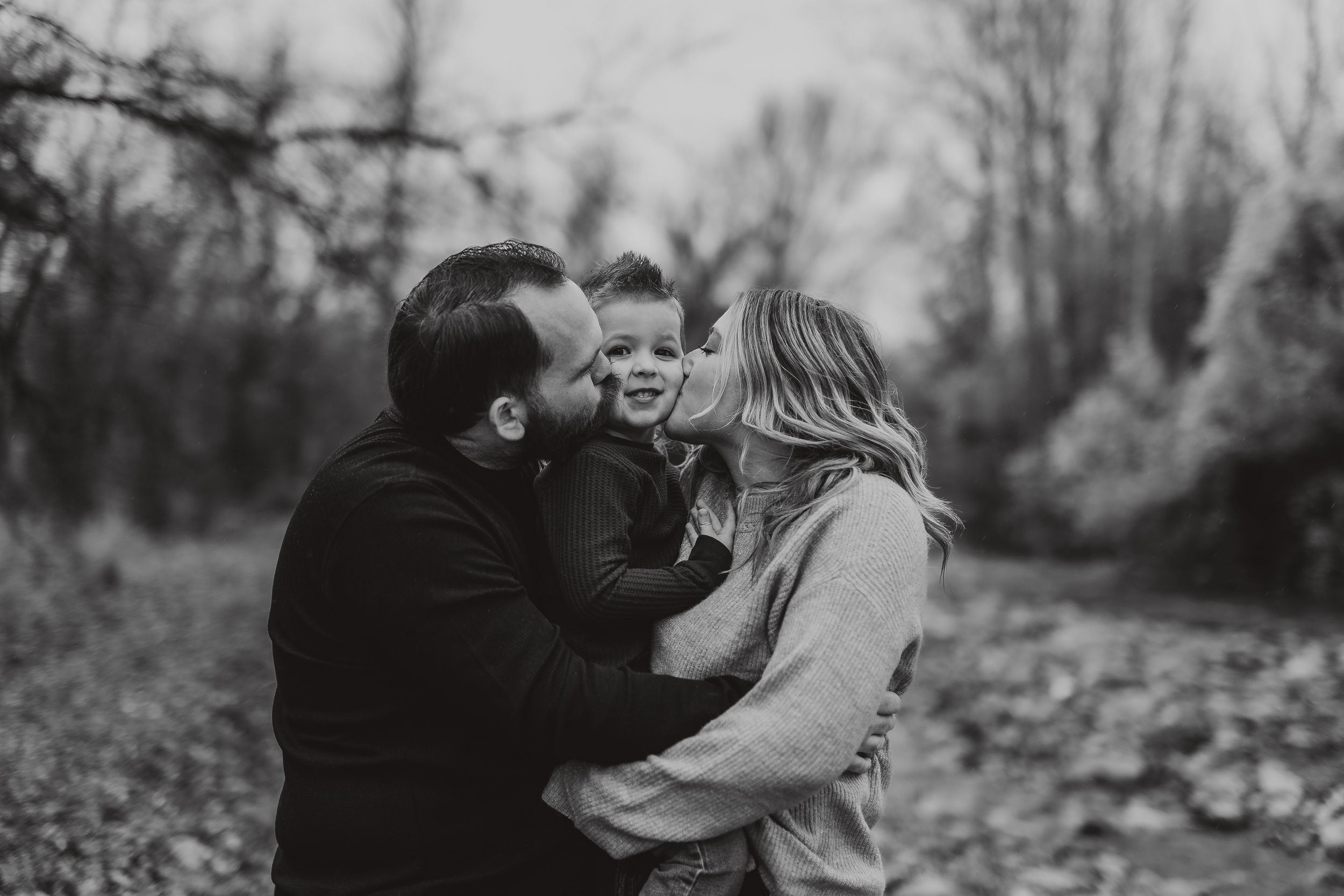 Black and white photo of parents kissing their young child on the cheeks outdoors in a wooded setting.