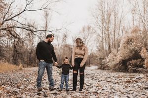Family of three holding hands on a rocky riverbank surrounded by bare autumn trees.
