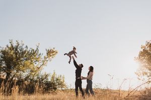 A joyful family plays outdoors as a father tosses a baby in the air while mother watches in a sunny field.