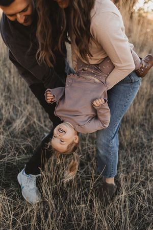 Parents playfully hold toddler upside down while laughing outdoors in a golden dry grass field during fall.