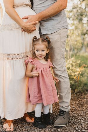 A toddler girl in a pink dress stands between her pregnant mother and father outdoors during a family maternity photo session.