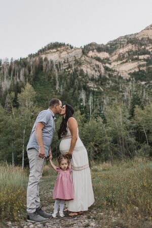 Pregnant woman in white dress kissing partner outdoors with toddler girl in pink dress, mountain backdrop.