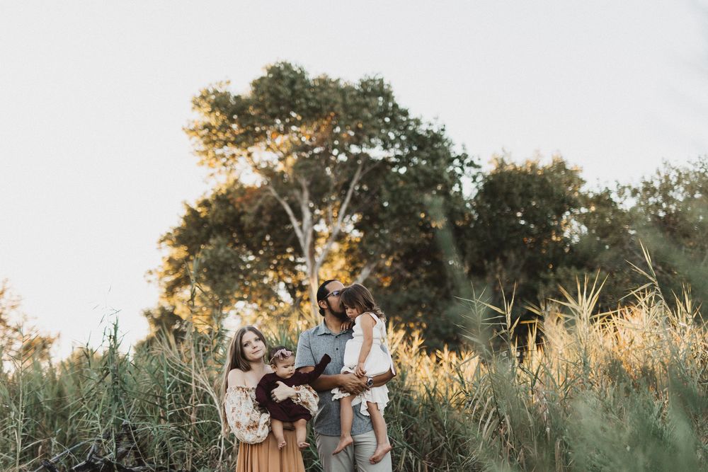 Family of four standing together in golden hour sunlight among tall green grasses and trees outdoors.