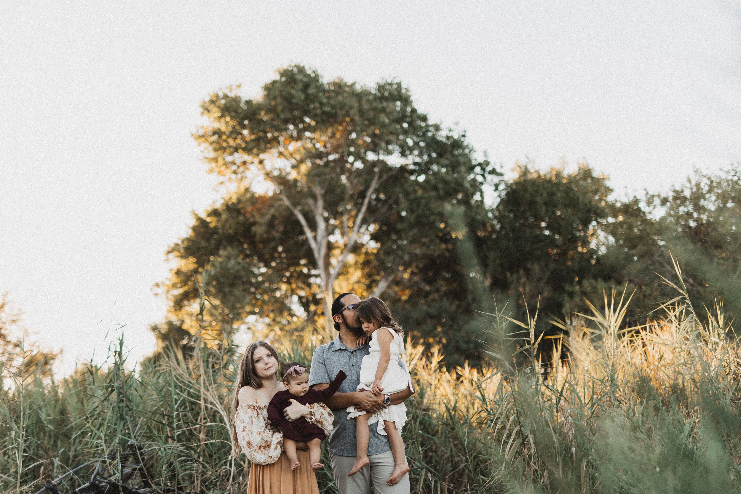 Family of four standing together in golden hour sunlight among tall green grasses and trees outdoors.