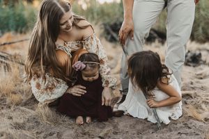 Mother sitting on sandy ground with two young daughters and a dog during an outdoor family photo session.