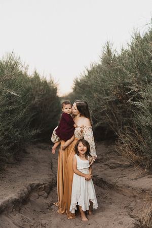 A mother in a golden skirt kisses her baby while her young daughter in a white dress smiles on a sandy path.
