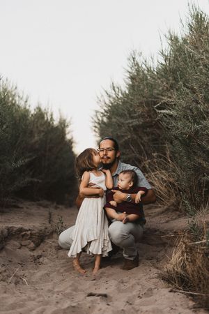 A father kneels on sandy ground holding a baby while a young girl kisses his cheek, surrounded by green shrubs.