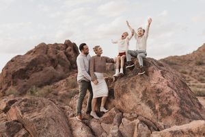 Family of four posing on rocky desert boulders, two adults standing and two children sitting with arms raised joyfully.