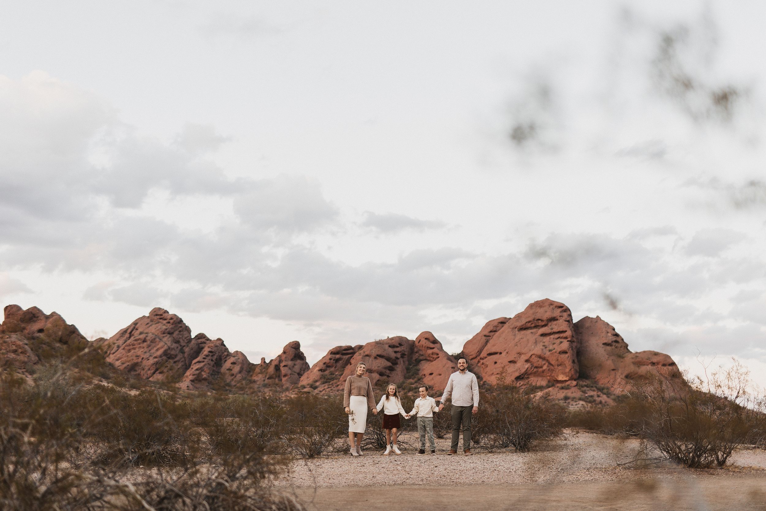 Family holding hands in desert landscape with red rock formations under cloudy sky.