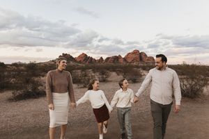 Family of four holding hands walking in desert landscape with red rock formations at sunset.
