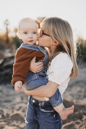 A young girl with glasses kisses a baby dressed in denim and brown sweater outdoors at golden hour.