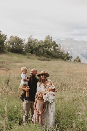 Family of four posing in a green meadow with mountain views, parents holding young children in boho-style outdoor portrait.