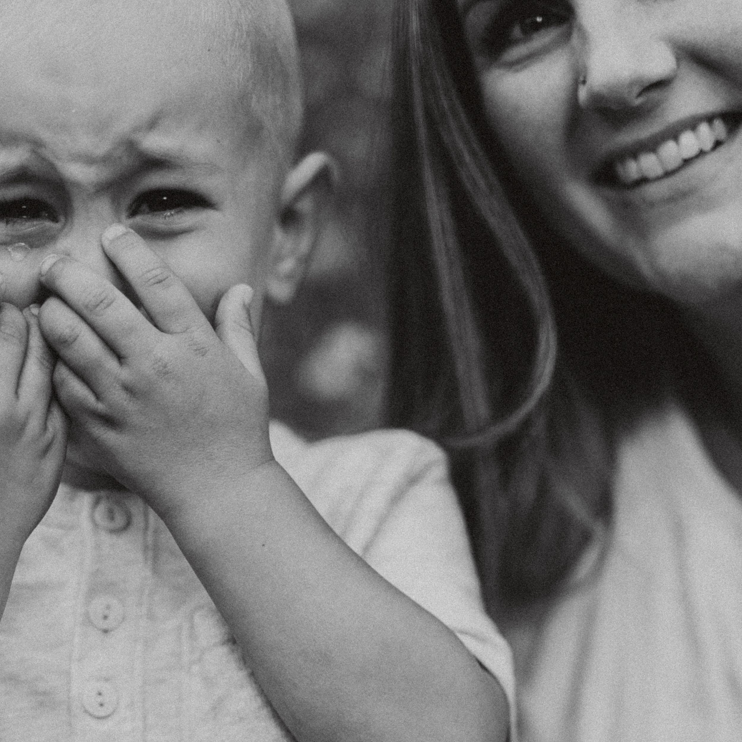 Black and white photo of a laughing woman holding a crying toddler covering their face with their hand.