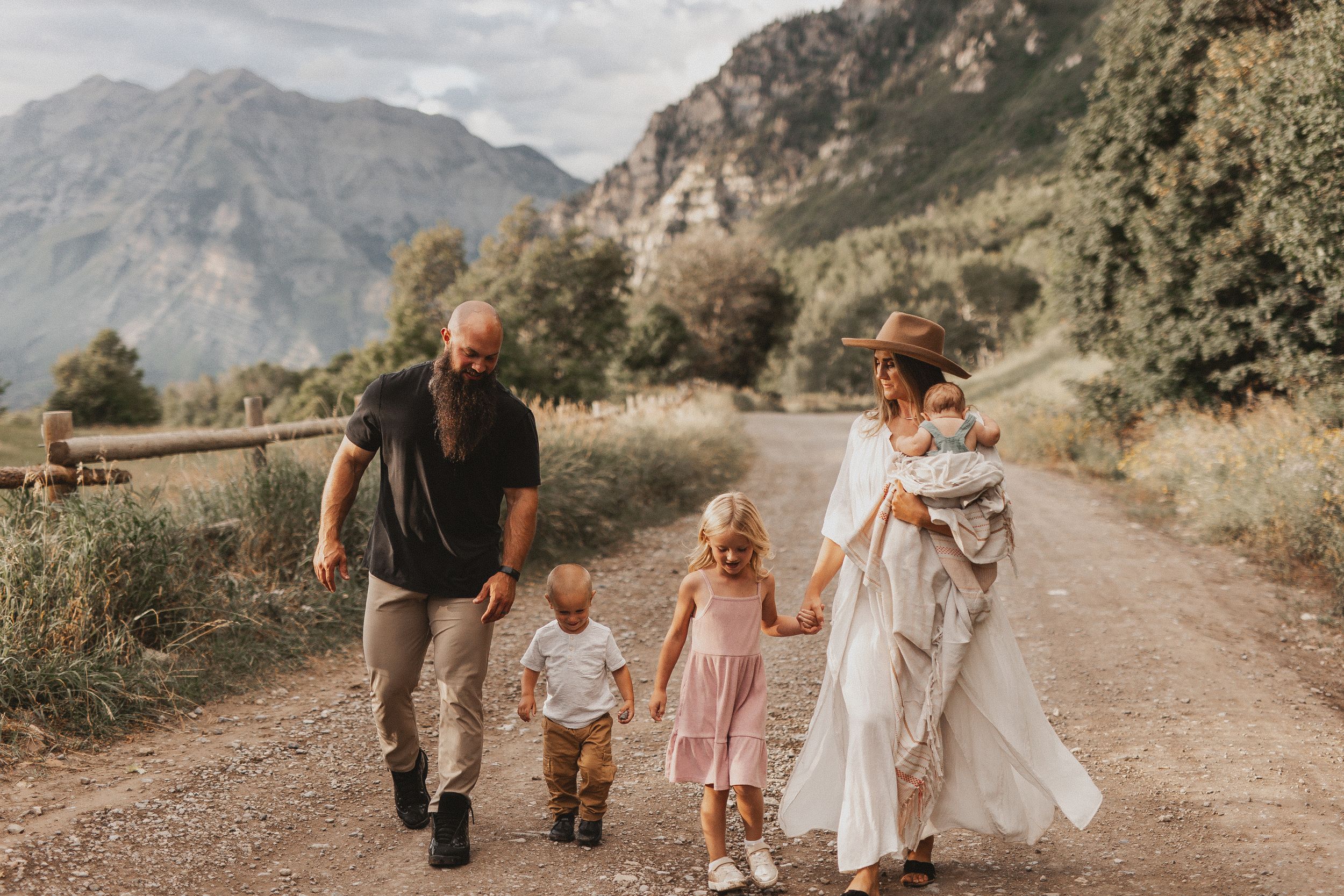 Family walking on a mountain dirt path, parents holding hands with young children, scenic peaks in the background.