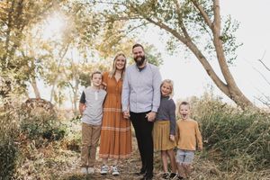 Family of five posing outdoors in coordinated fall outfits among golden trees in warm sunlight.