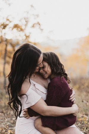 A mother and daughter share a joyful embrace outdoors during autumn, both smiling with eyes closed.