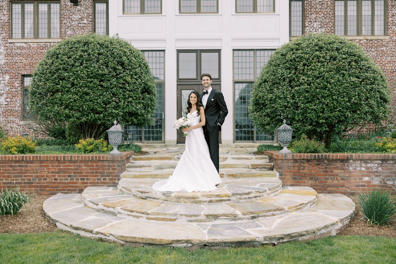 Bride and groom pose on stone steps outside elegant brick mansion with manicured hedges on their wedding day.