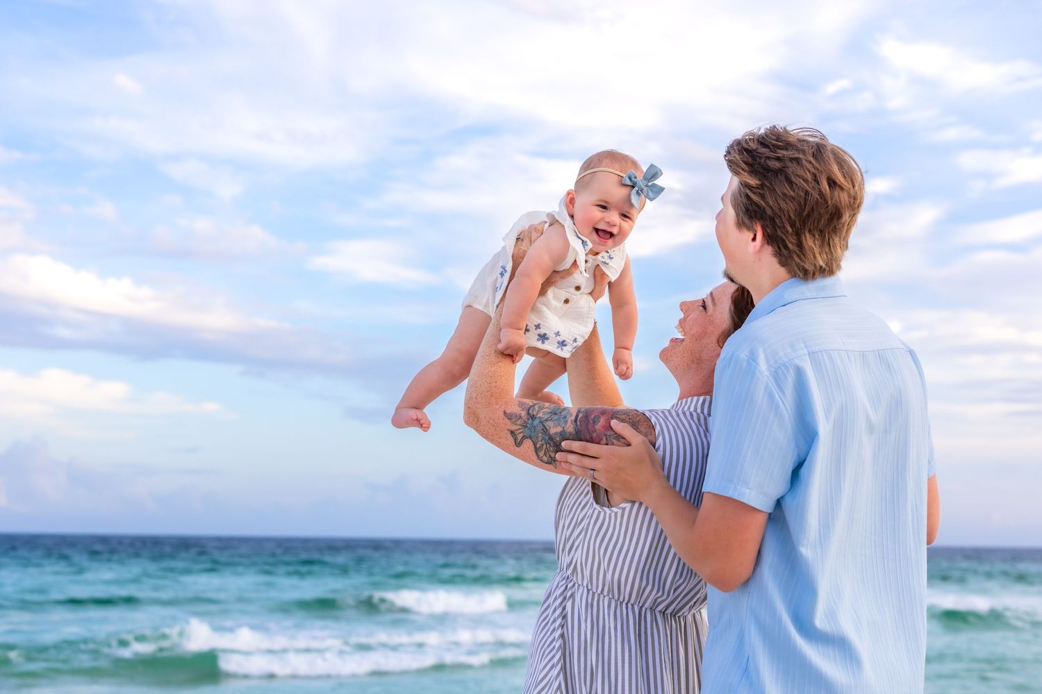 Happy family at beach, parents lifting laughing baby girl in air with ocean waves and cloudy sky background.