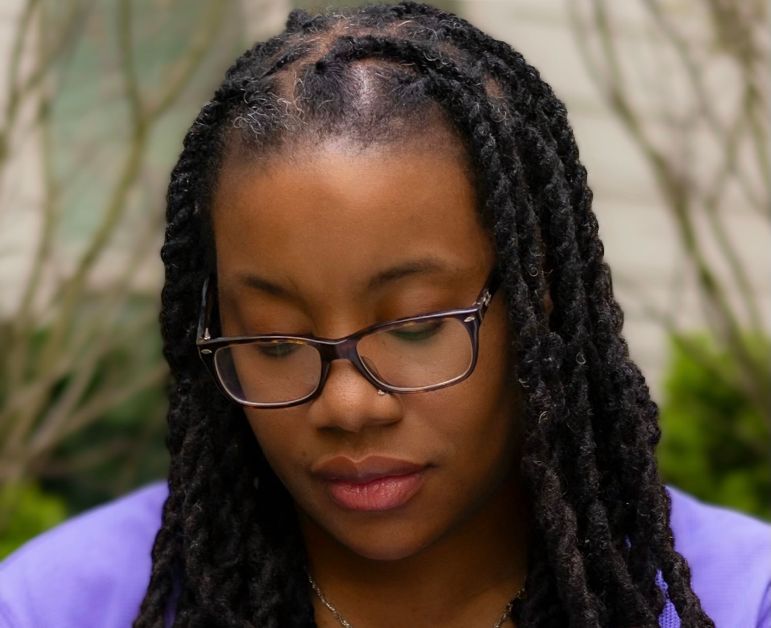 Young Black woman with braided locs and glasses looking downward in a contemplative pose outdoors.