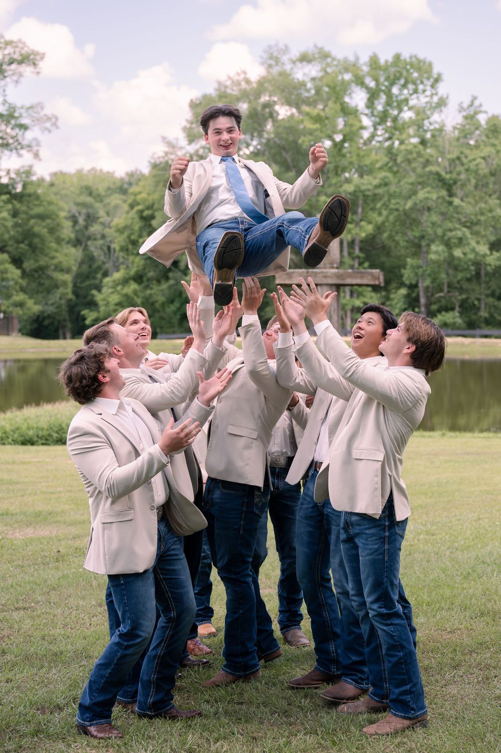Groomsmen tossing groom in the air outdoors near a pond, all wearing beige blazers and jeans.