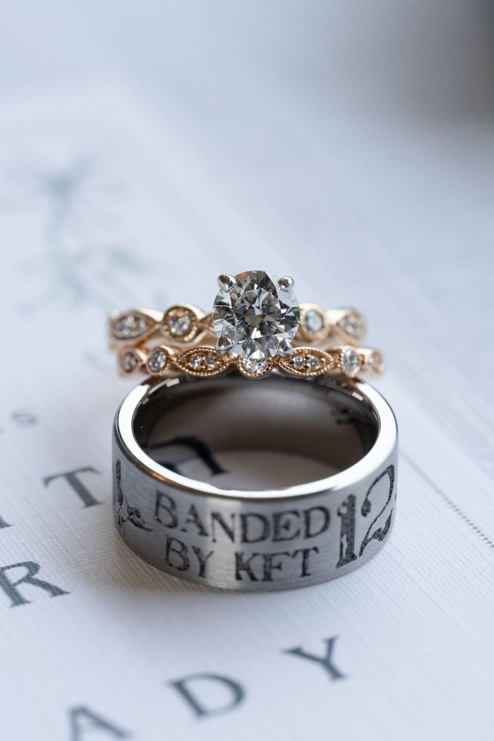 Gold diamond engagement ring stacked with engraved tungsten wedding band reading 'Banded by KFT' on white book.