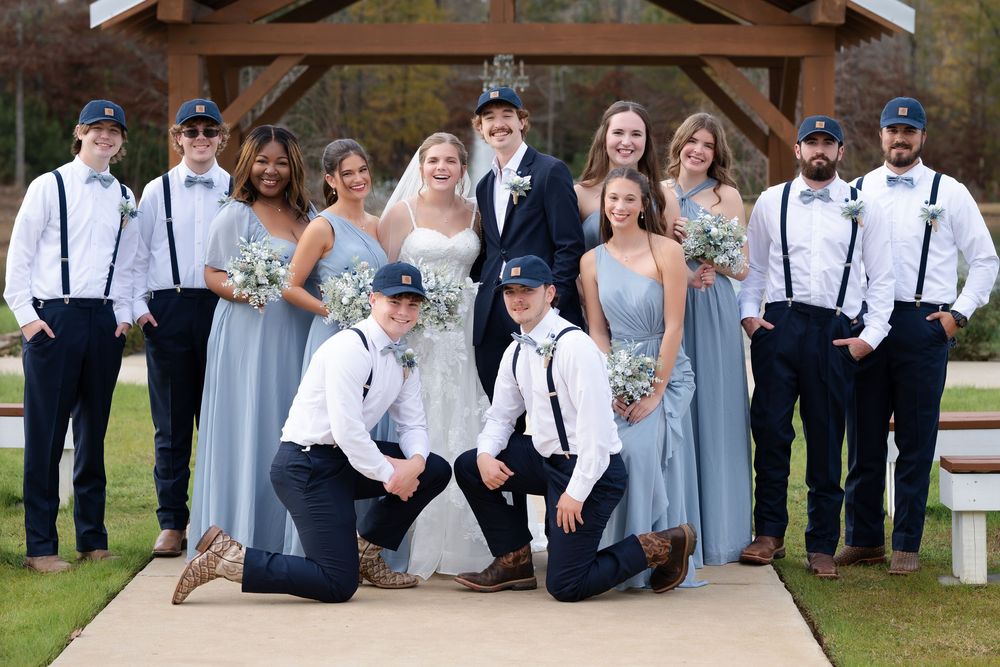 Wedding party poses outdoors in blue bridesmaid dresses and suspenders with bow ties under a wooden gazebo.