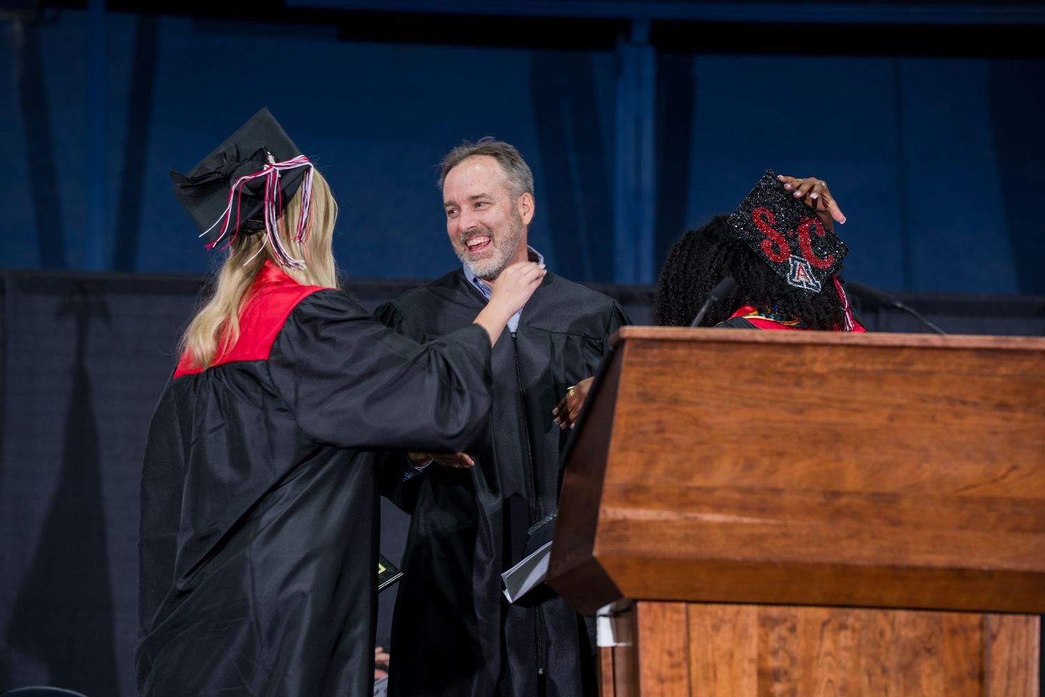 Two graduates in black robes shake hands on stage at a graduation ceremony near a wooden podium.