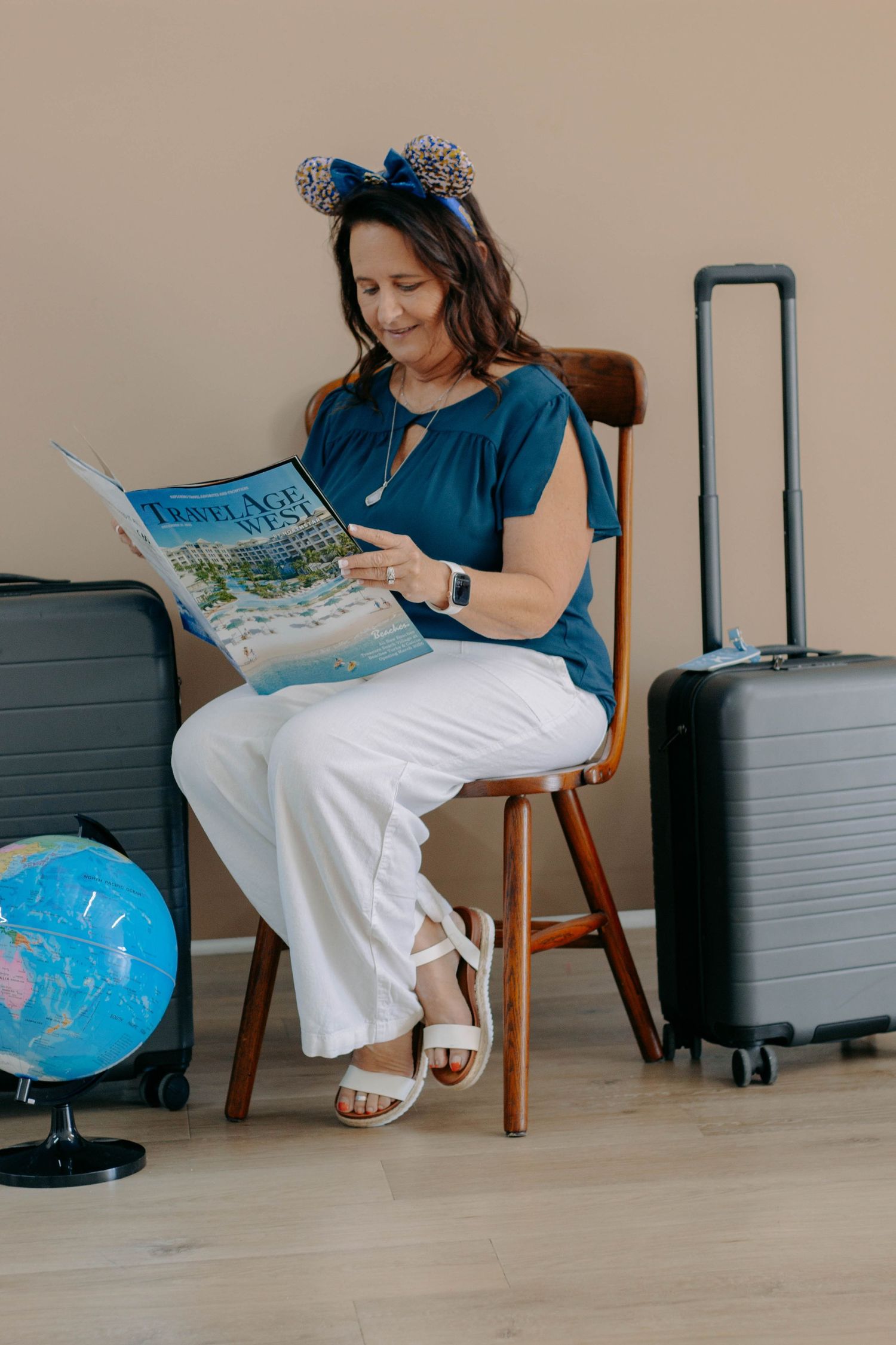 Woman in blue top and white pants wearing Mickey ears reads a travel magazine while seated between suitcases and a globe.