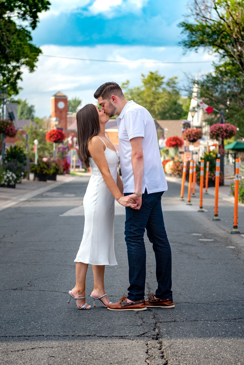 Couple sharing a kiss on a scenic street lined with colorful hanging flower baskets and a clock tower.
