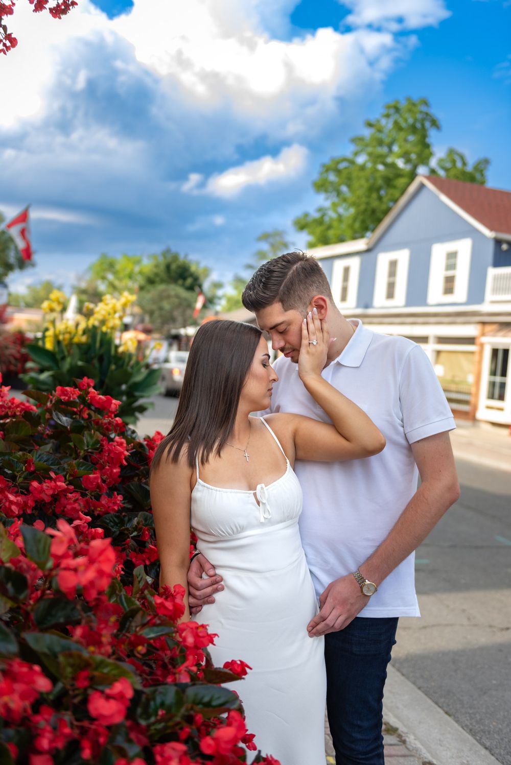 Romantic couple nearly kissing beside vibrant red flowers under a blue cloudy sky.