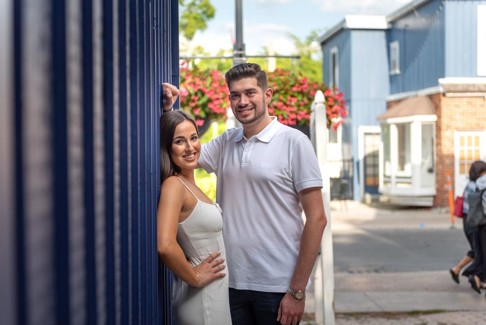 Smiling couple peeking around a blue building corner with red flowers behind them.